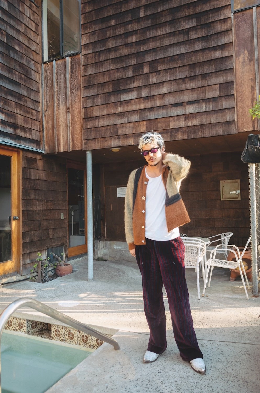 A man with bleached hair and tinted sunglasses in a brown and cream mohair cardigan, white tank top and dark striped trousers with silver cowboy boots, standing beside a pool in a cedar-shingled courtyard.
