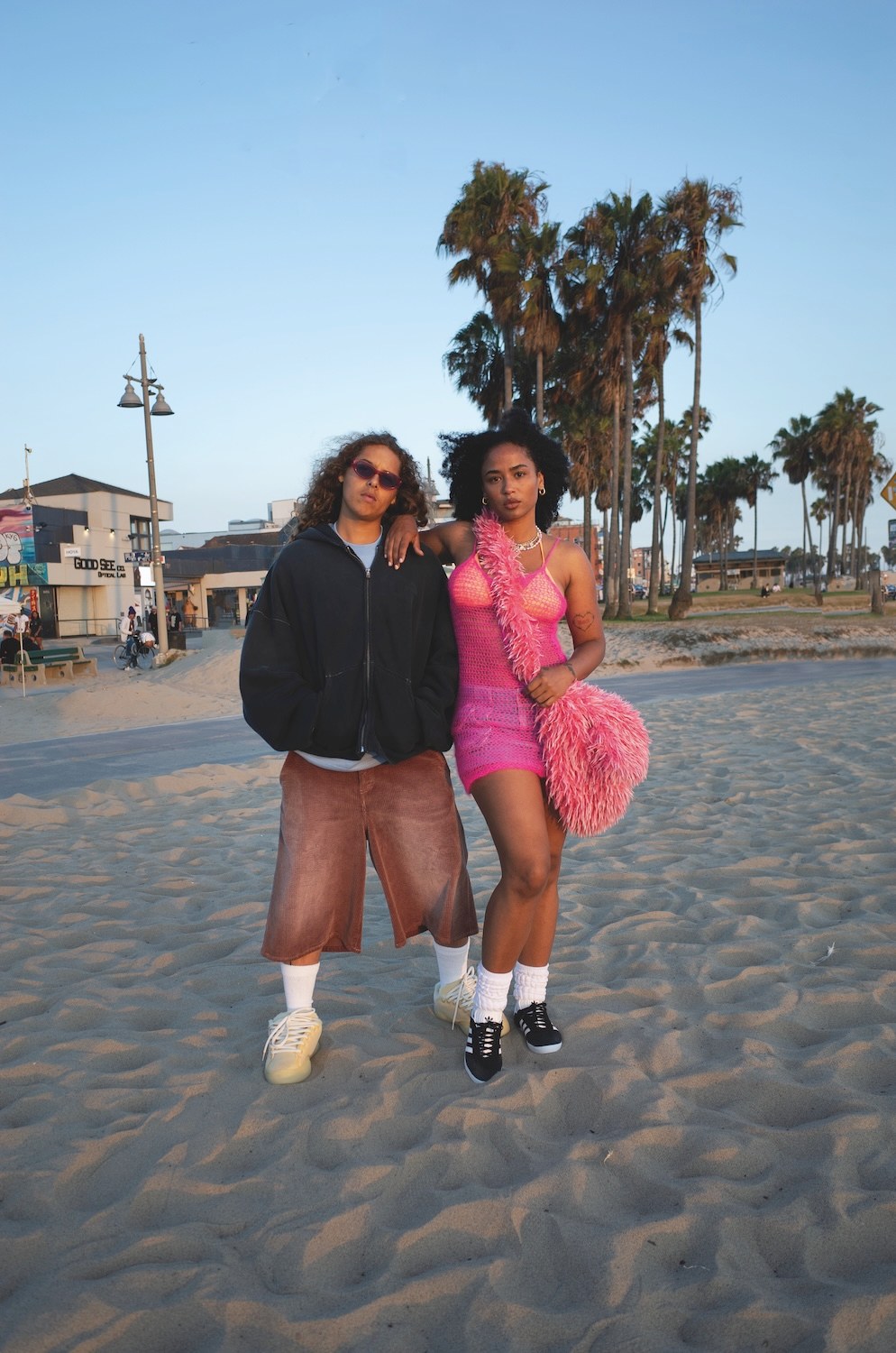 Two people standing together on the sand at sunset, one in an oversized black zip hoodie and brown corduroy shorts, the other in a bright pink crochet dress with a pink faux fur bag, palm trees behind them.