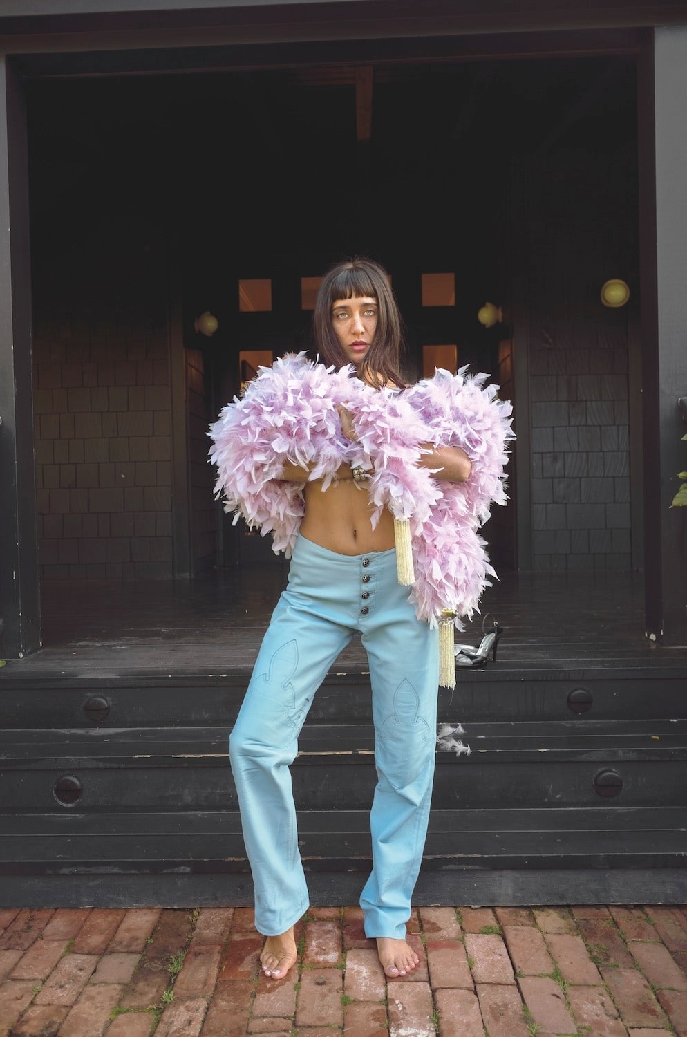 Jesse Jo Stark wrapped in a lilac feather stole over pale blue jeans, standing barefoot on a brick path in front of a dark-painted house.